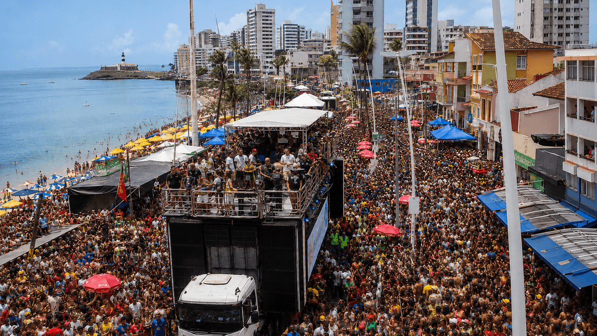 Un trio elétrico avanza entre la multitud del Carnaval de Salvador en la costa Barra-Ondina, con el Farol da Barra visible a lo lejos.